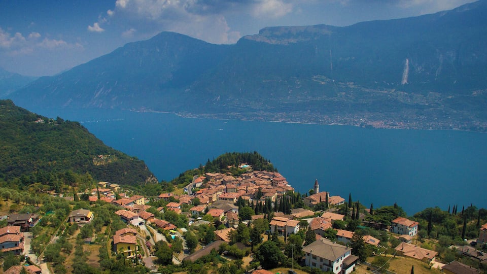 A coastal village with red-roofed houses, a mountain range, and a large body of water.