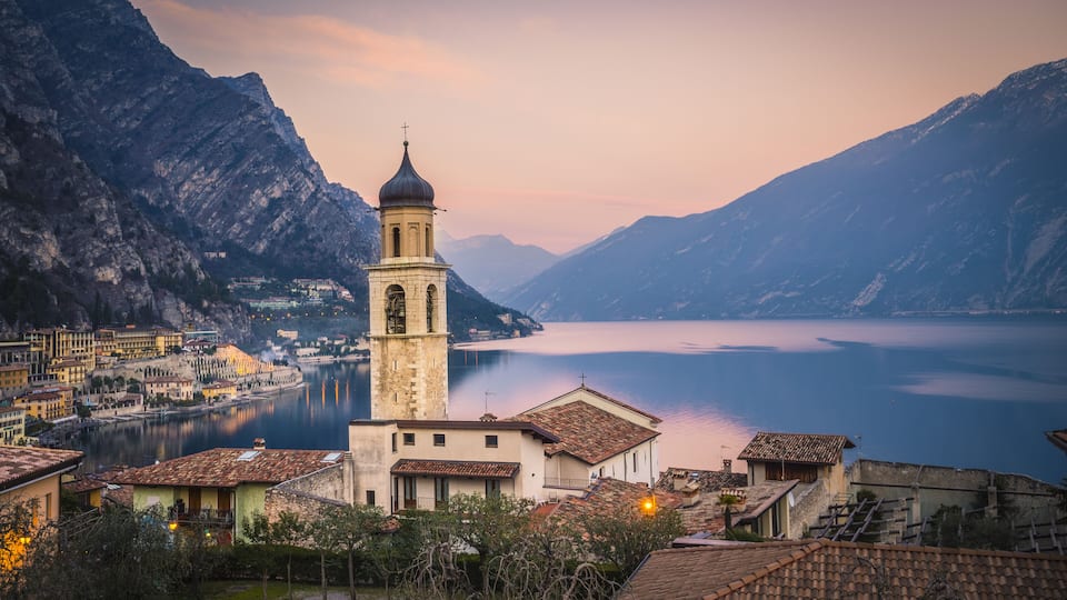 A historic church with a bell tower, a calm lake, and mountainous terrain in the background.