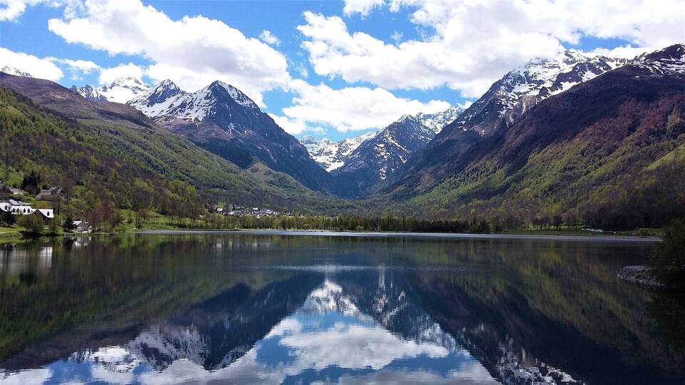 A mountain lake with reflections, surrounded by forested hills and snow-capped peaks.