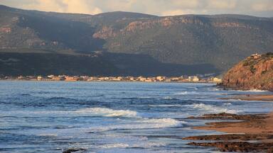 A coastal town with a rocky shoreline, waves crashing onto the beach, and mountains in the background.