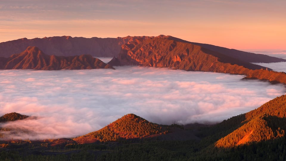 Uma cordilheira com um pico arborizado e um vale sob uma camada de nuvens.