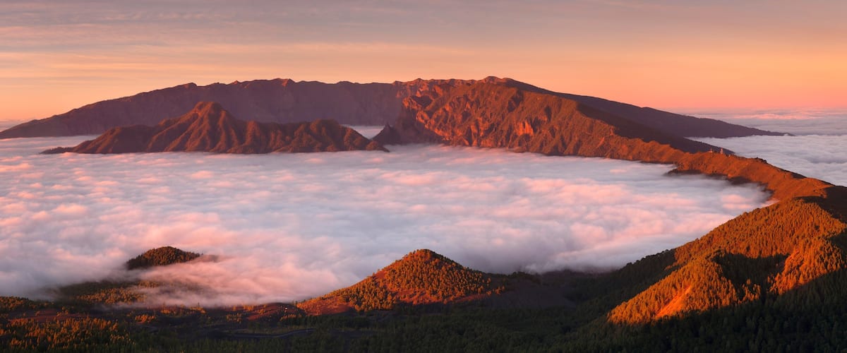 A mountain range with a forested peak and a valley below a cloud layer.