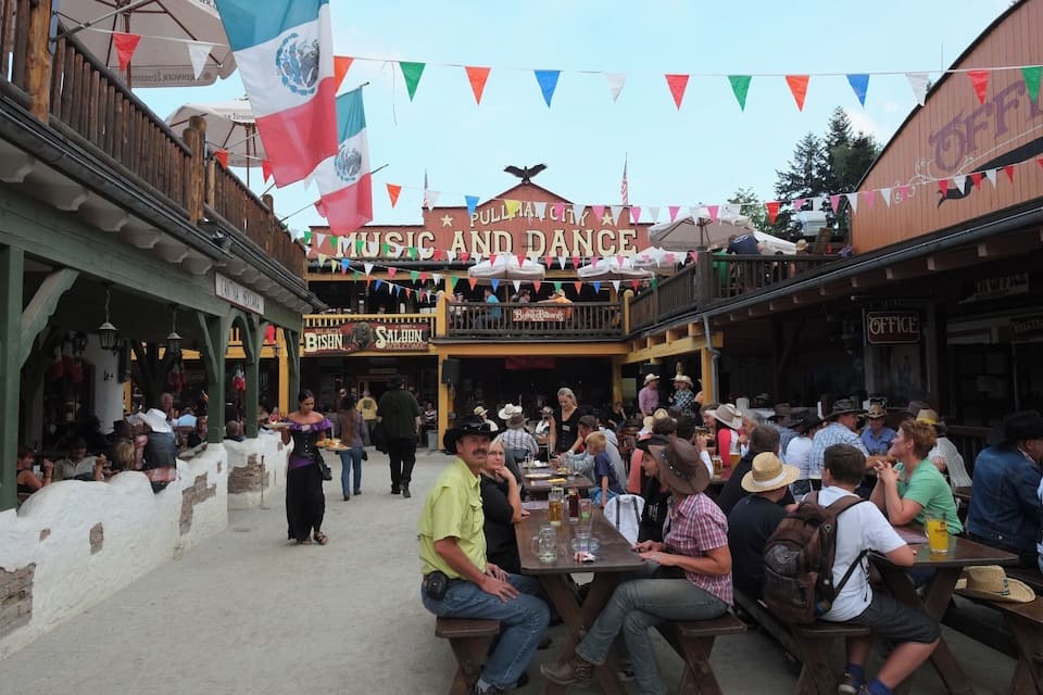 A bustling outdoor dining area with a 'PULLMAN CITY MUSIC AND DANCE' sign, surrounded by colorful flags and a crowd of people.