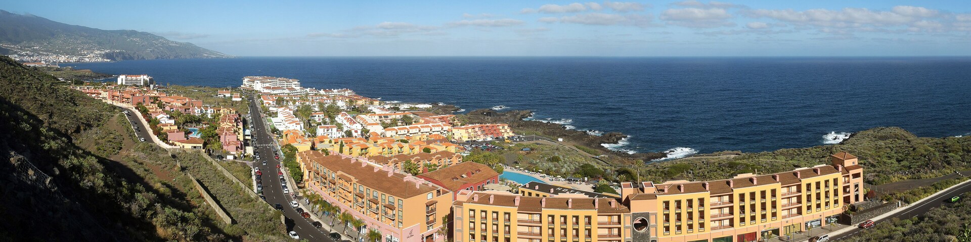 A coastal town with buildings along the sea, a road, and a mountainous backdrop.