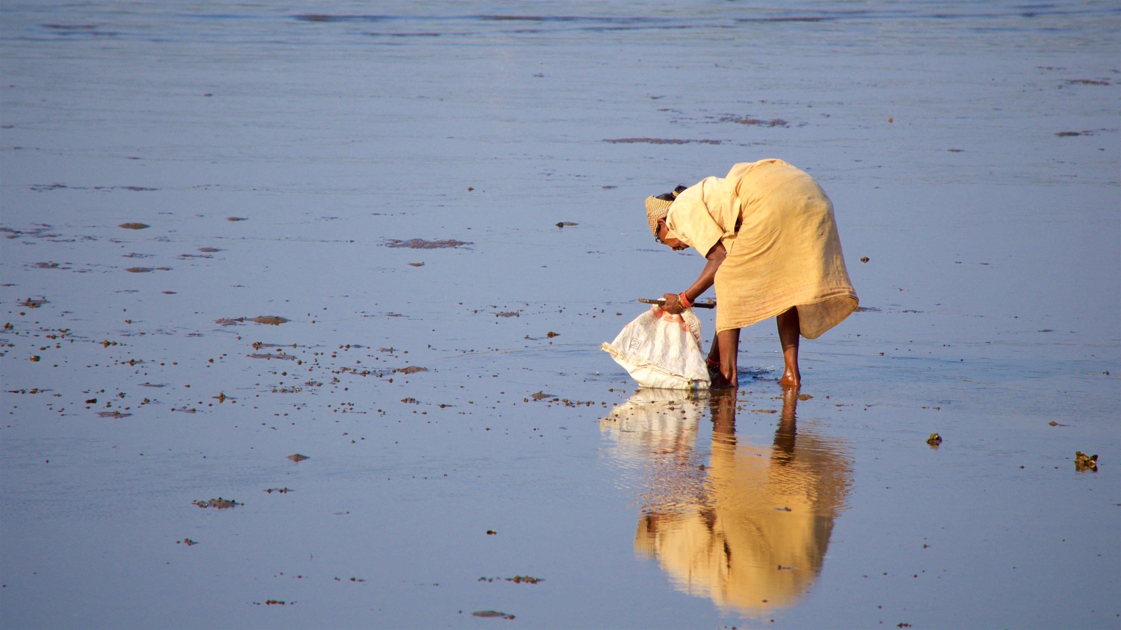 A person in a yellow dress bending over to pick up a white bag from a sandy beach with scattered debris.