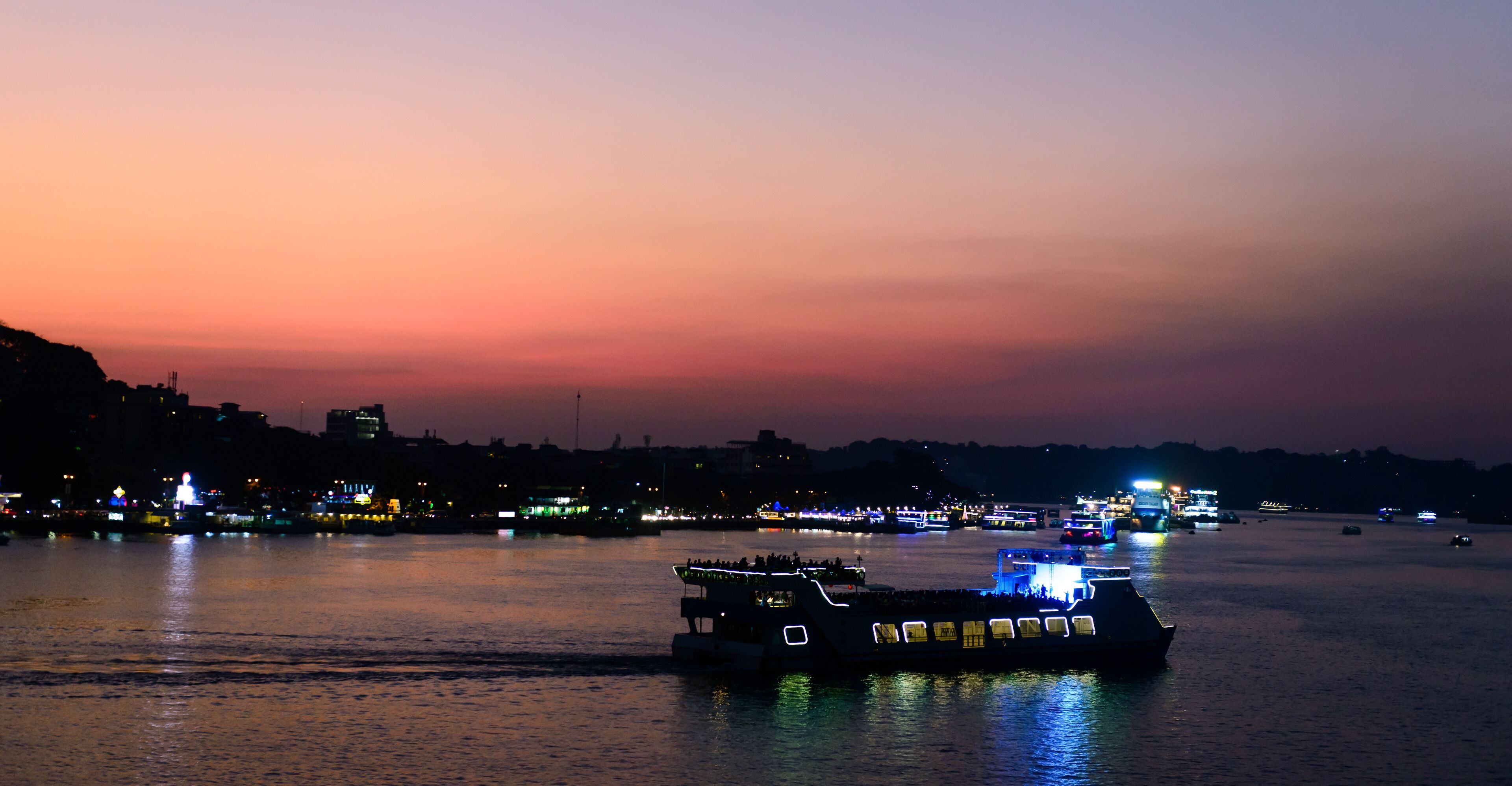A river at sunset with illuminated boats and a city skyline in the background.