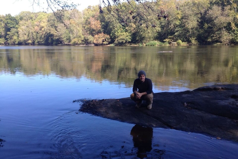 A person sitting on a rock by a calm river surrounded by trees.