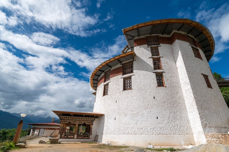 A white stone tower with a curved roof and wooden accents, surrounded by mountains and a clear sky.