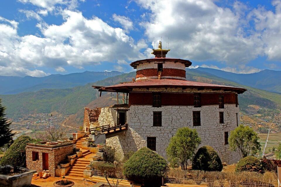 A traditional Tibetan prayer wheel building with a red roof and white walls, surrounded by lush greenery and mountains in the background.