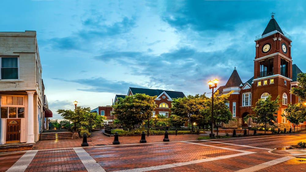 A historic town square with a prominent clock tower, brick buildings, and a well-maintained tree-lined street.