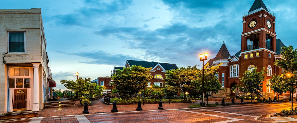 A historic town square with a prominent clock tower, brick buildings, and a well-maintained tree-lined street.