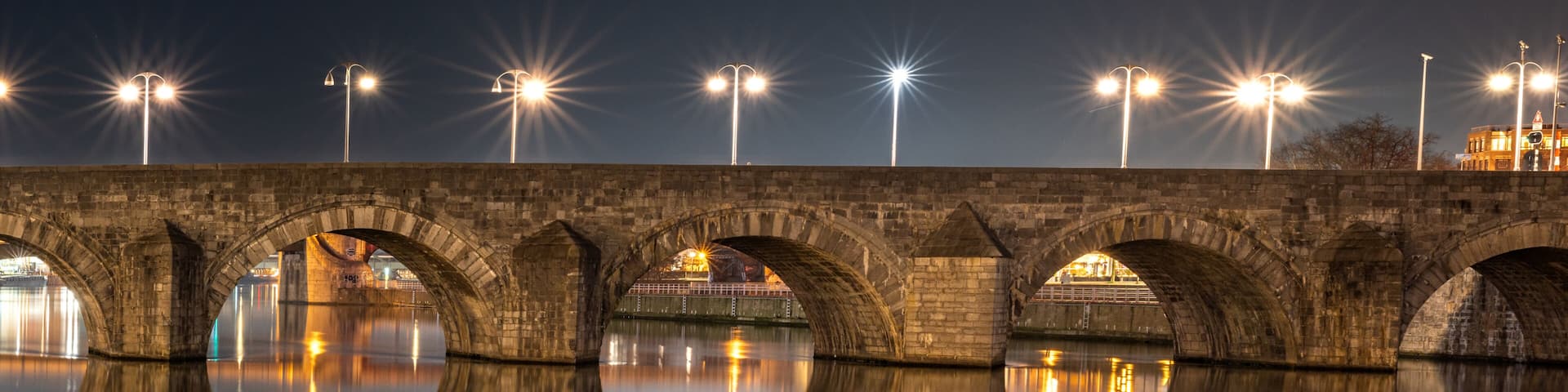 A stone bridge with multiple arches over a calm body of water, reflecting the bridge and street lights.