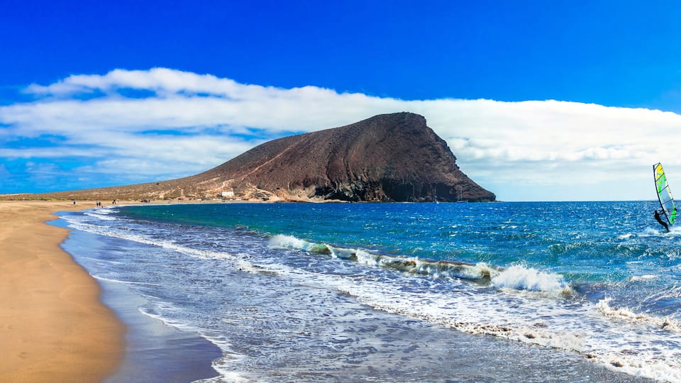 A beach with a large mountain in the background and a person windsurfing.