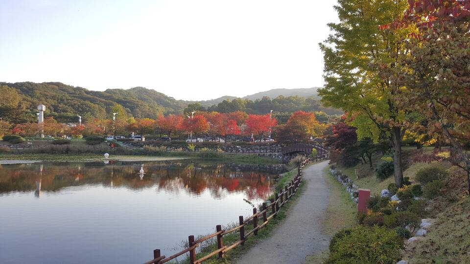 A serene lakeside path with autumn foliage, a wooden bridge, and distant hills.