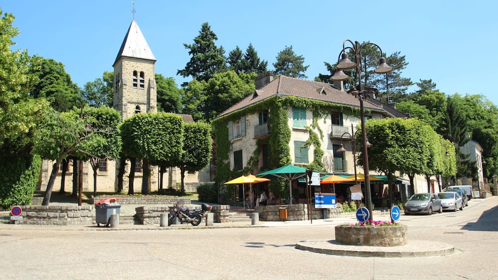 Une place de ville avec un clocher d’église, un bâtiment recouvert de lierre et des voitures garées.