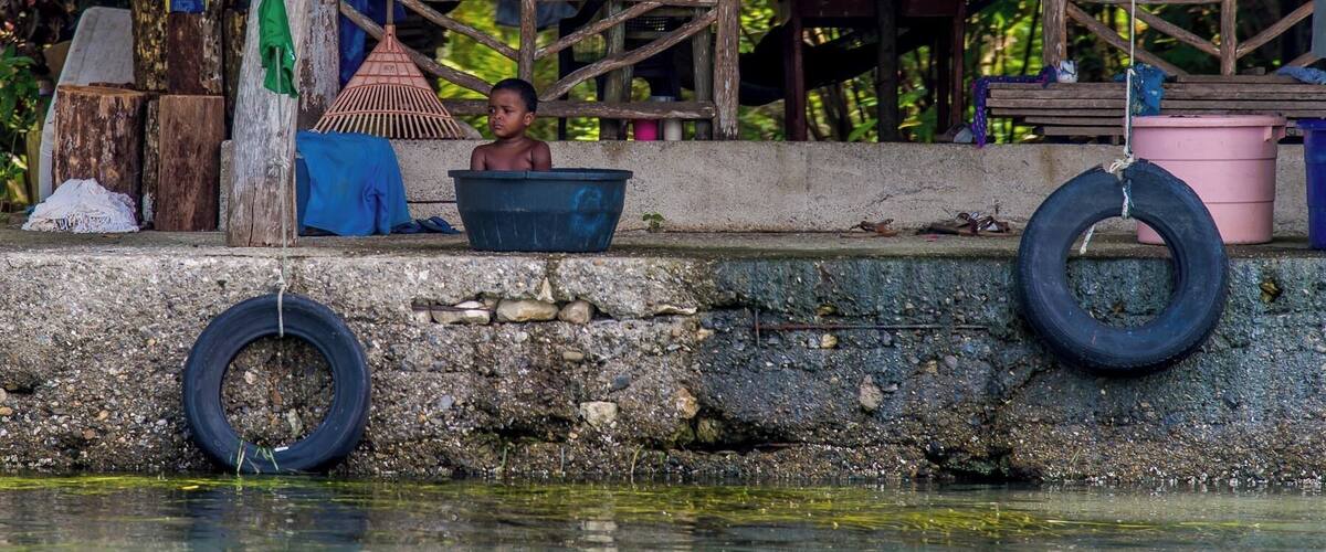 A rural scene with a child sitting in a basin, surrounded by wooden structures and a water body.