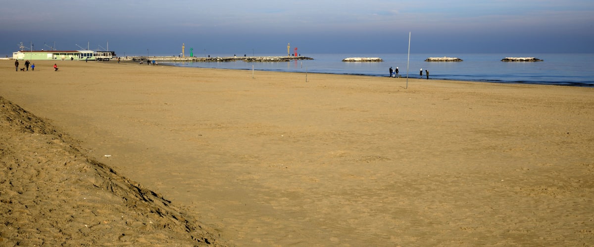A wide sandy beach with a few people, a pier, and a lighthouse in the distance.