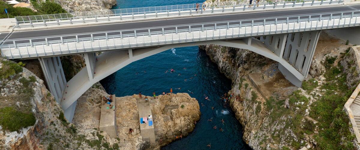 A bridge over a rocky coastline with people swimming in the water below.