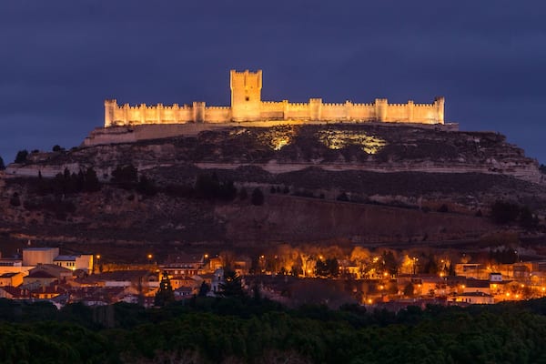 A medieval castle atop a hill with a town below, surrounded by hills.