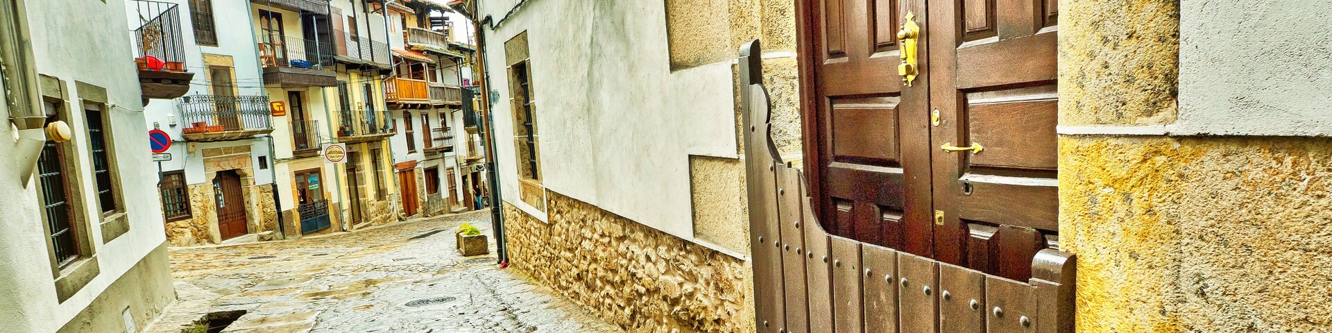 A narrow cobblestone street flanked by buildings with balconies and a large wooden door.