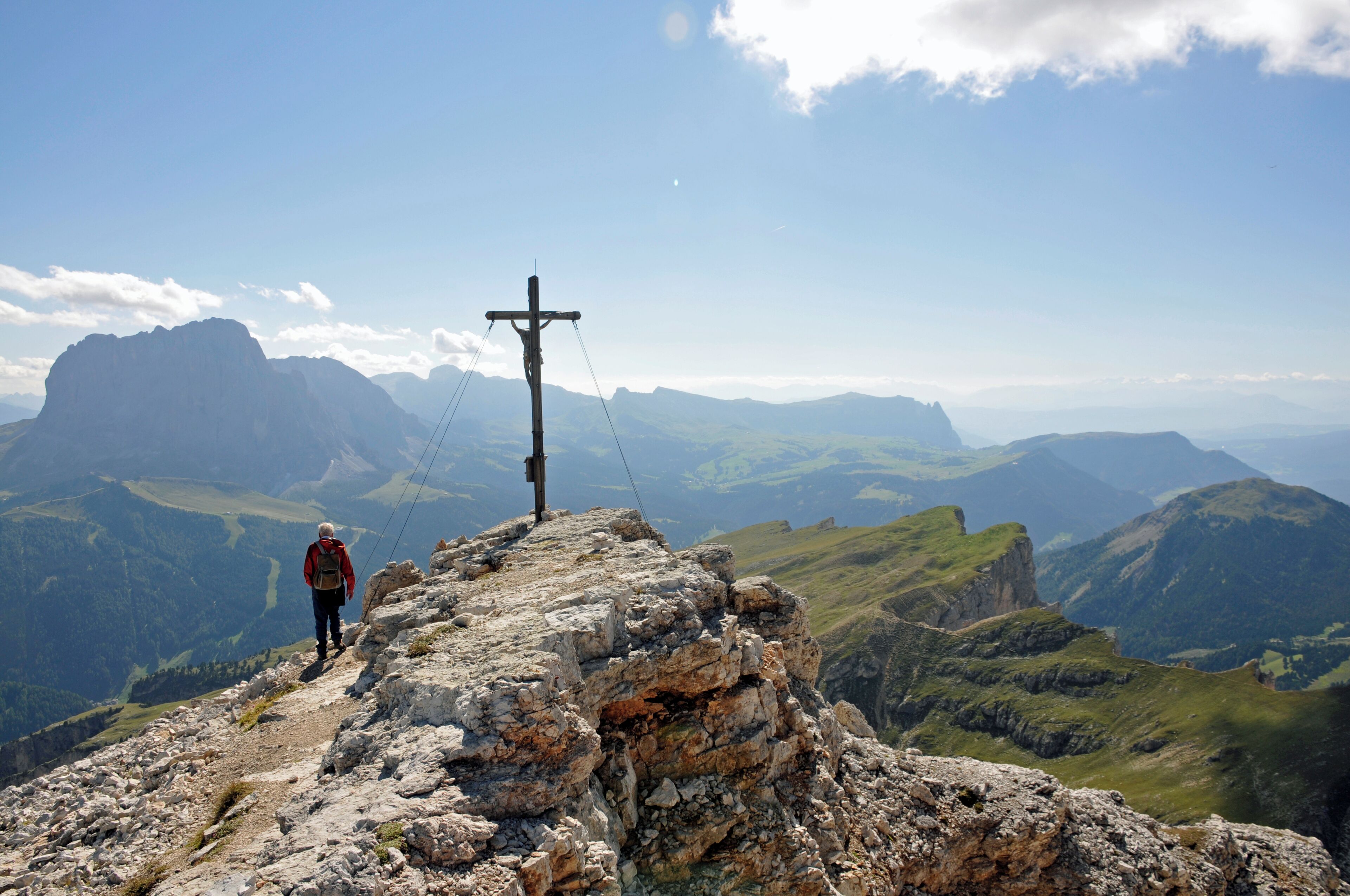 A hiker on a rocky mountain path with a cross in the distance and a vast mountainous landscape.