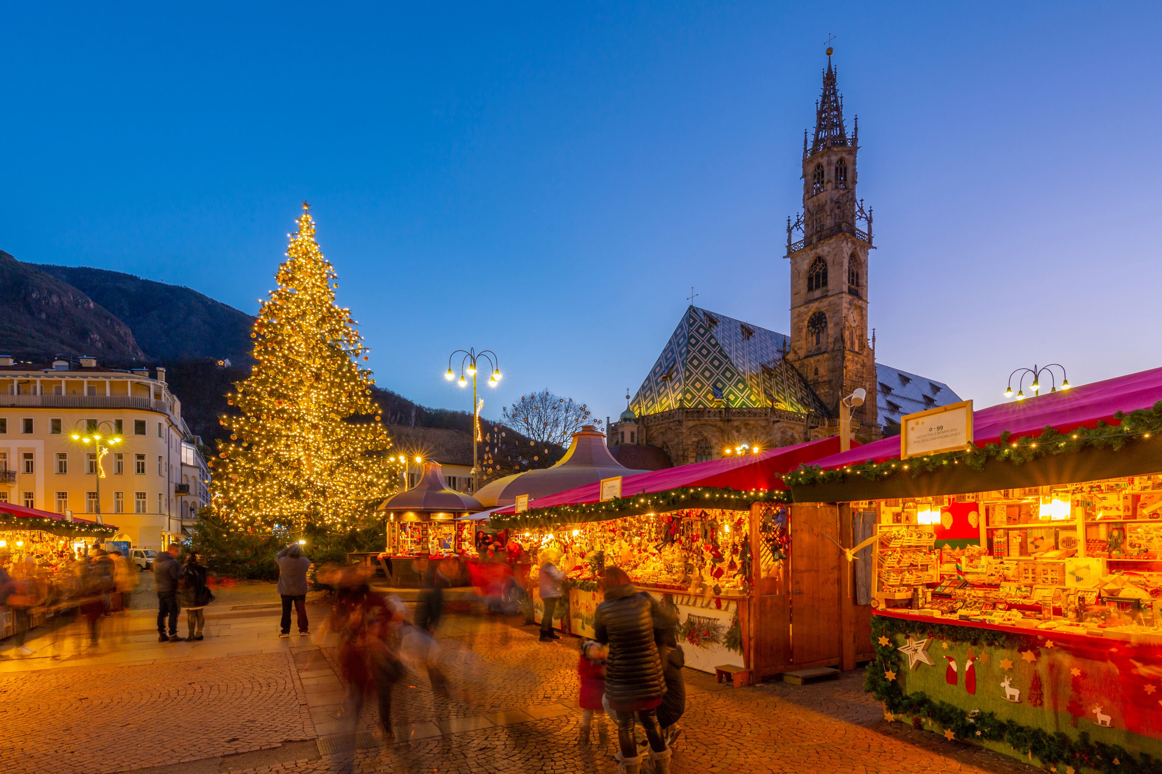 Um mercado de Natal com uma grande árvore iluminada e uma torre de igreja histórica ao fundo.