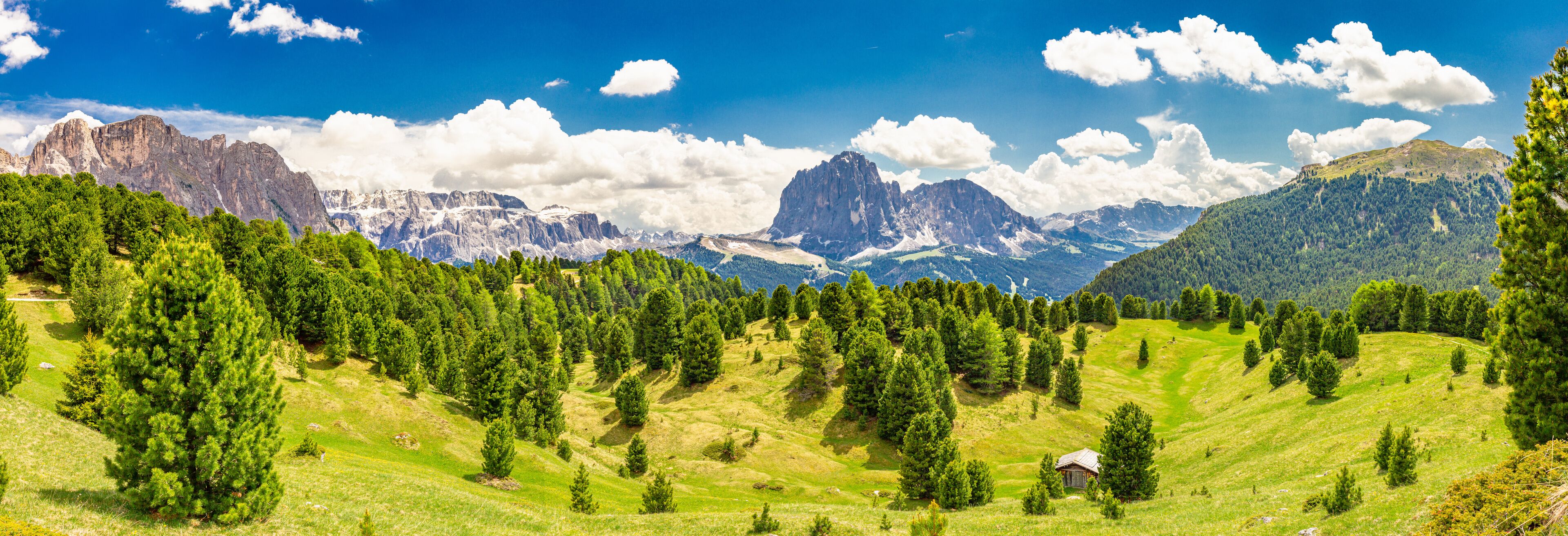 A mountainous landscape with dense forests, a small cabin, and prominent peaks under a clear blue sky.