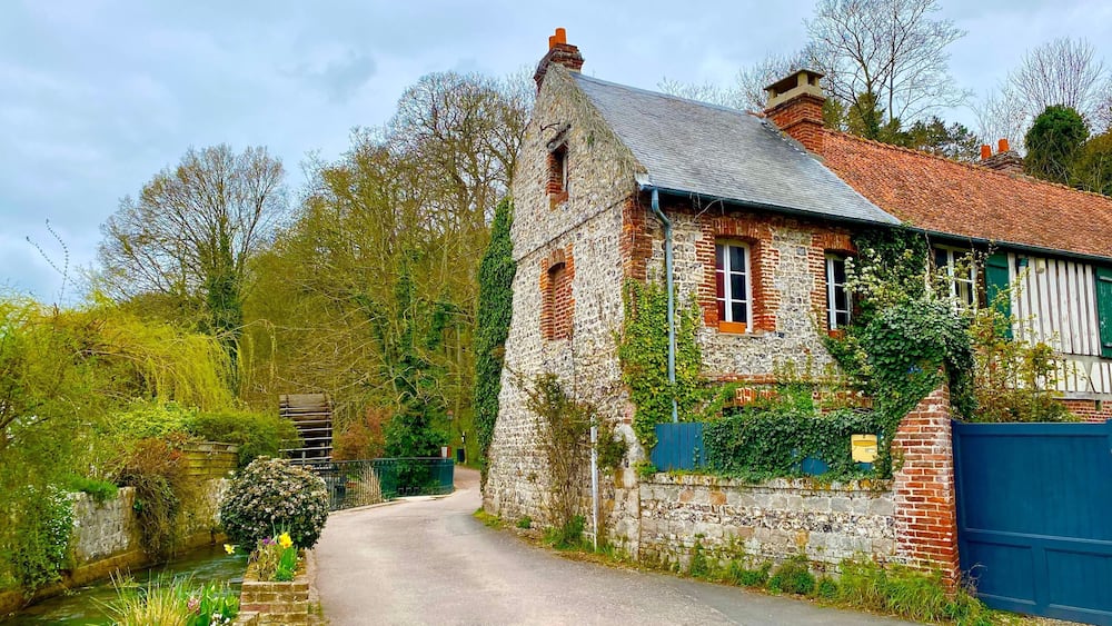 Une maison en pierre avec un toit recouvert de tuiles rouges, entourée de verdure et bordée d’une étroite route.