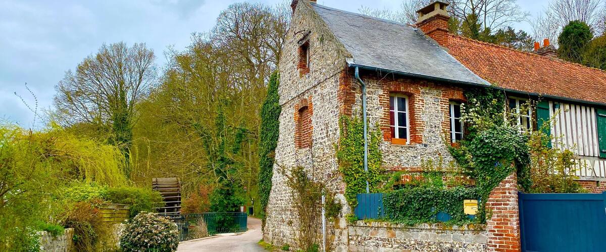 A stone house with a red-tiled roof, surrounded by greenery and a narrow road.