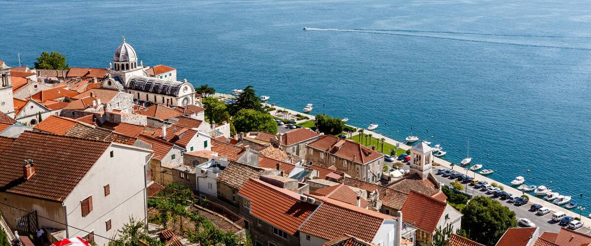 A coastal town with terracotta-roofed buildings, a prominent church with a dome, and a marina with boats.