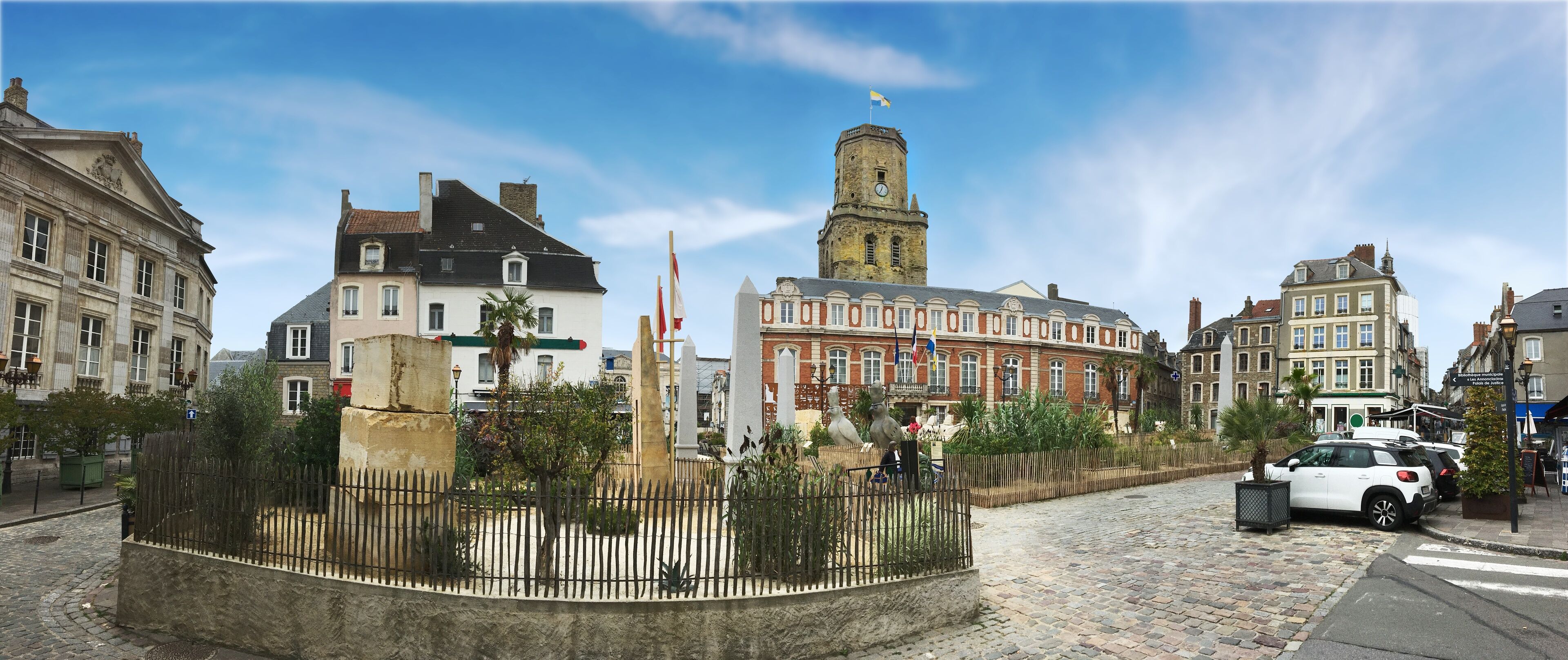 A cobblestone street with historic buildings, a central tower, and a fenced garden area.