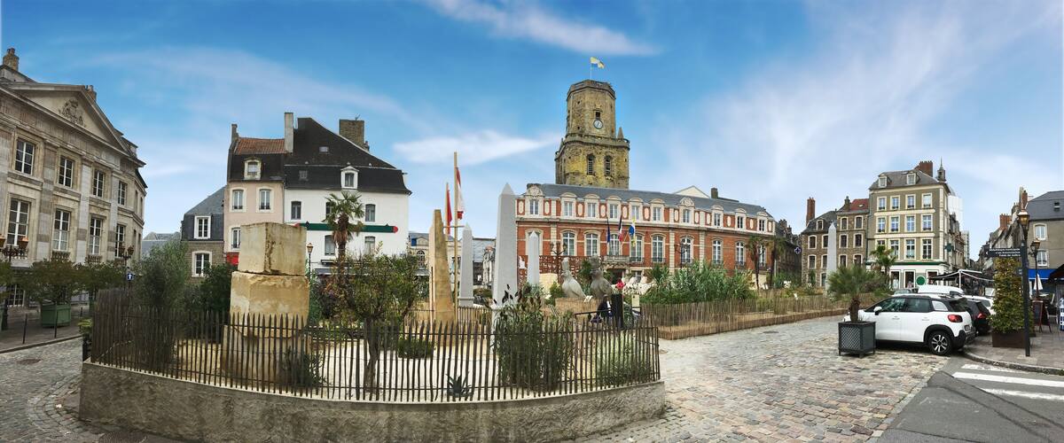 A cobblestone street with historic buildings, a central tower, and a fenced garden area.