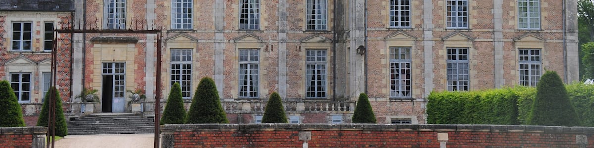 Un grand bâtiment historique, doté de nombreuses cheminées, d’une entrée imposante et d’un jardin bien entretenu avec des haies taillées.