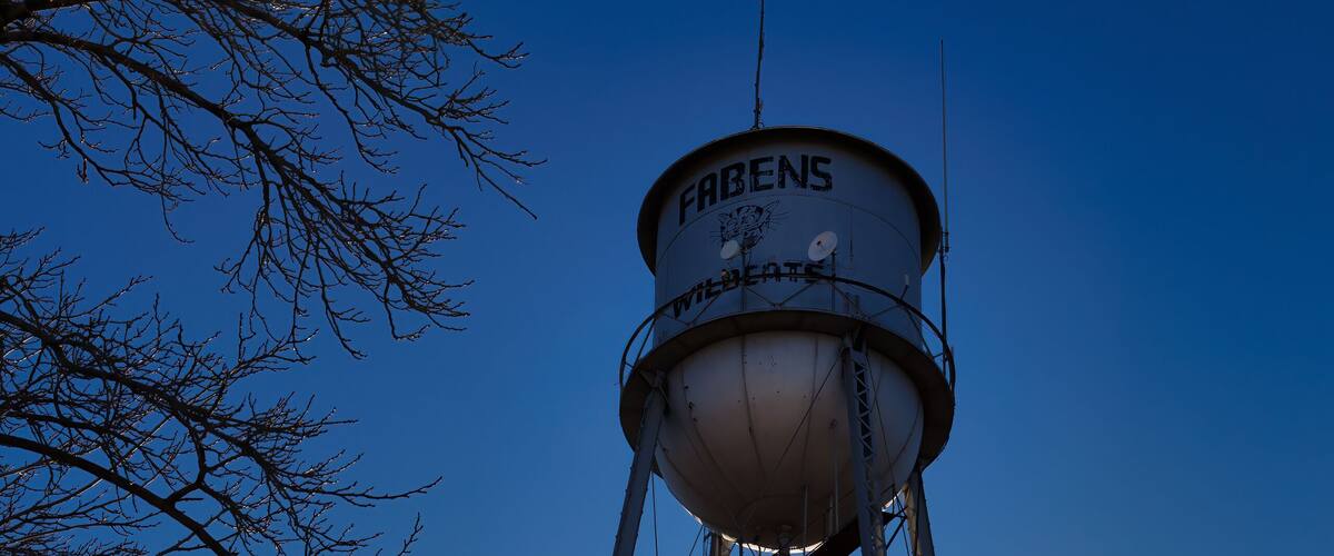 A water tower with the name "FABENS" and "WILDBURST" on it, set against a clear blue sky with the sun shining.