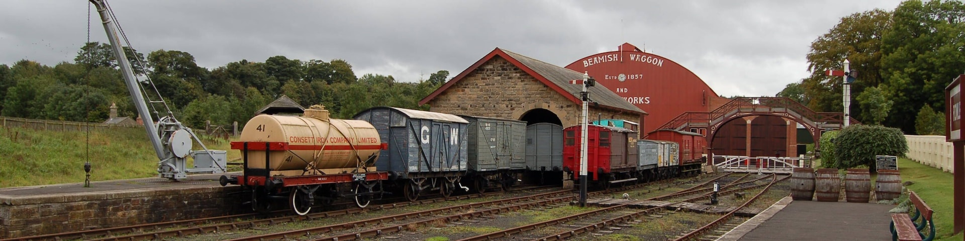 Una estación de tren con múltiples vías, un edificio de ladrillo rojo identificado como 'BERMISH WHOGON AND WORKS', y una grúa.