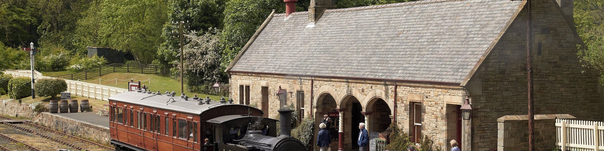 A historic train station with a steam locomotive, stone architecture, and a platform with people.