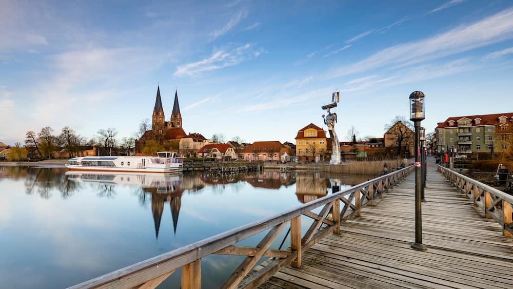 Ein ruhiger Fluss mit einer Holzbrücke, einem weißen Boot und einer Kirche mit zwei Türmen.