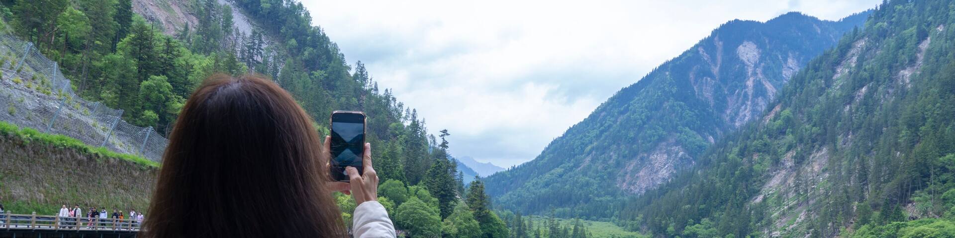 Asian female backpacker travels to Jiuzhaigou Valley in China, holding a smartphone to take pictures of the beautiful valley and lake reflection in the bright blue water.