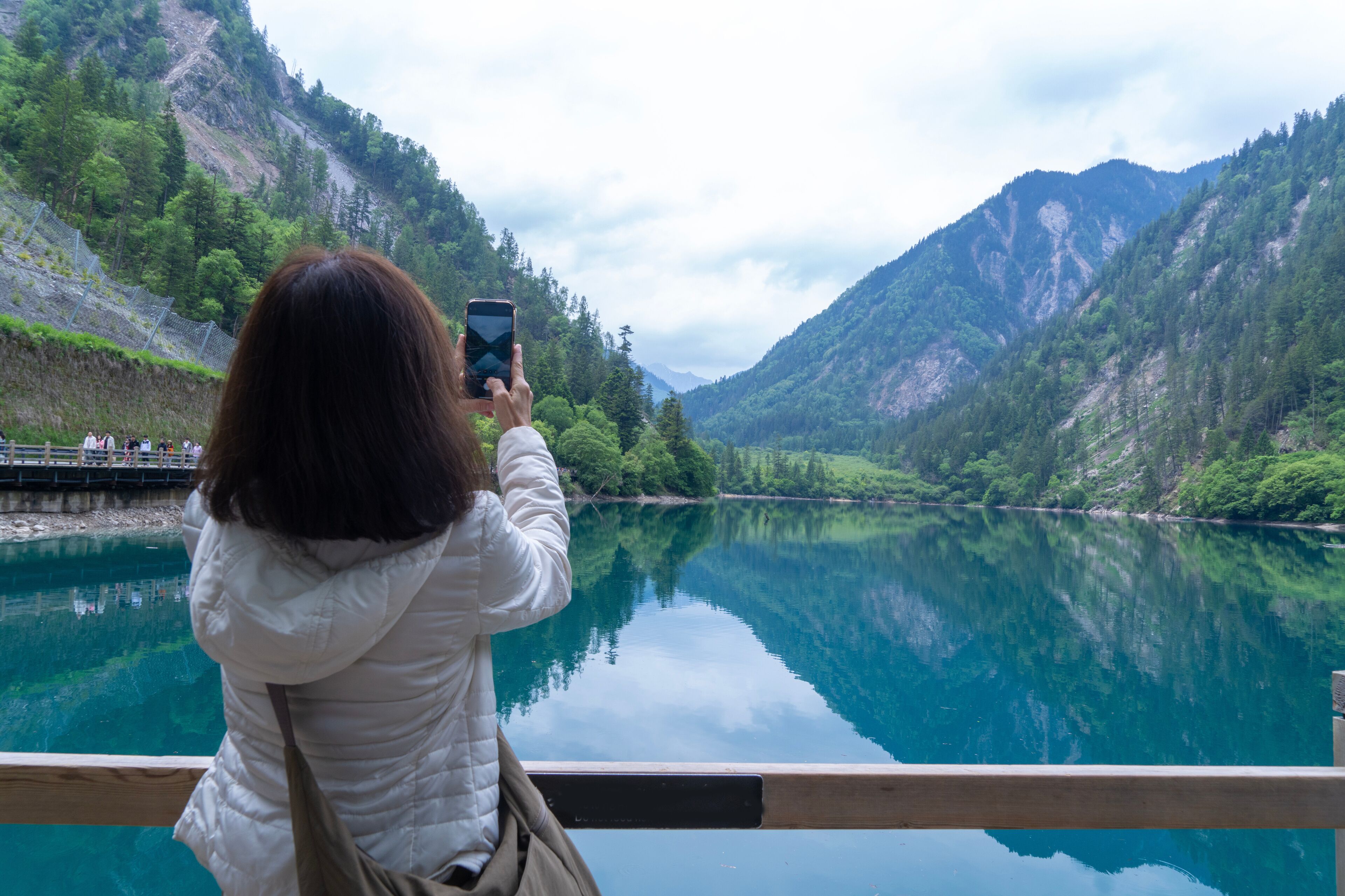 A woman taking a photo of a calm lake surrounded by mountains.