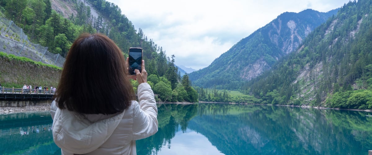 A woman taking a photo of a calm lake surrounded by mountains.