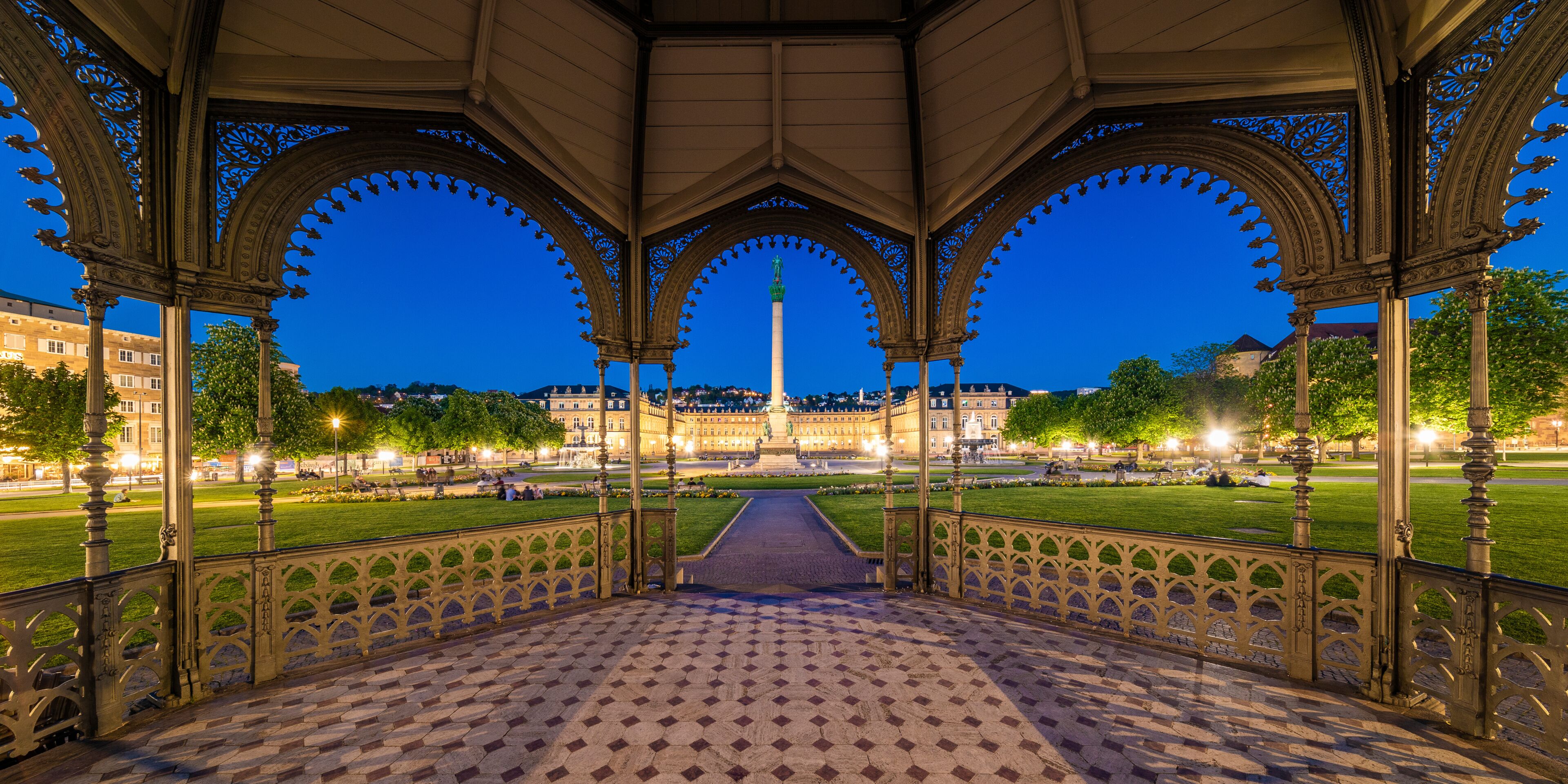 A nighttime view of a park with ornate arches, a central monument, and a well-lit pathway.