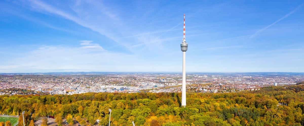 Ein hoher Turm mit einer Flagge, umgeben von Bäumen in Herbstfarben und einer Stadtkulisse im Hintergrund.