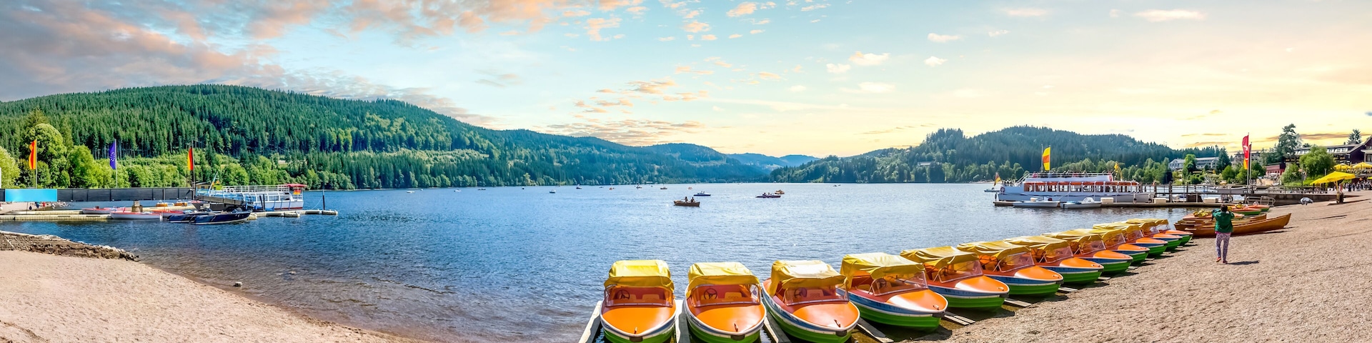 A lakeside with boats, a dock, and a forested mountain in the background.