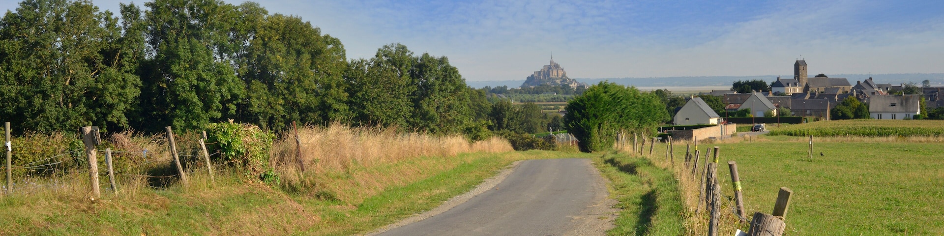 A rural road flanked by fields and trees, leading to a distant historic building.