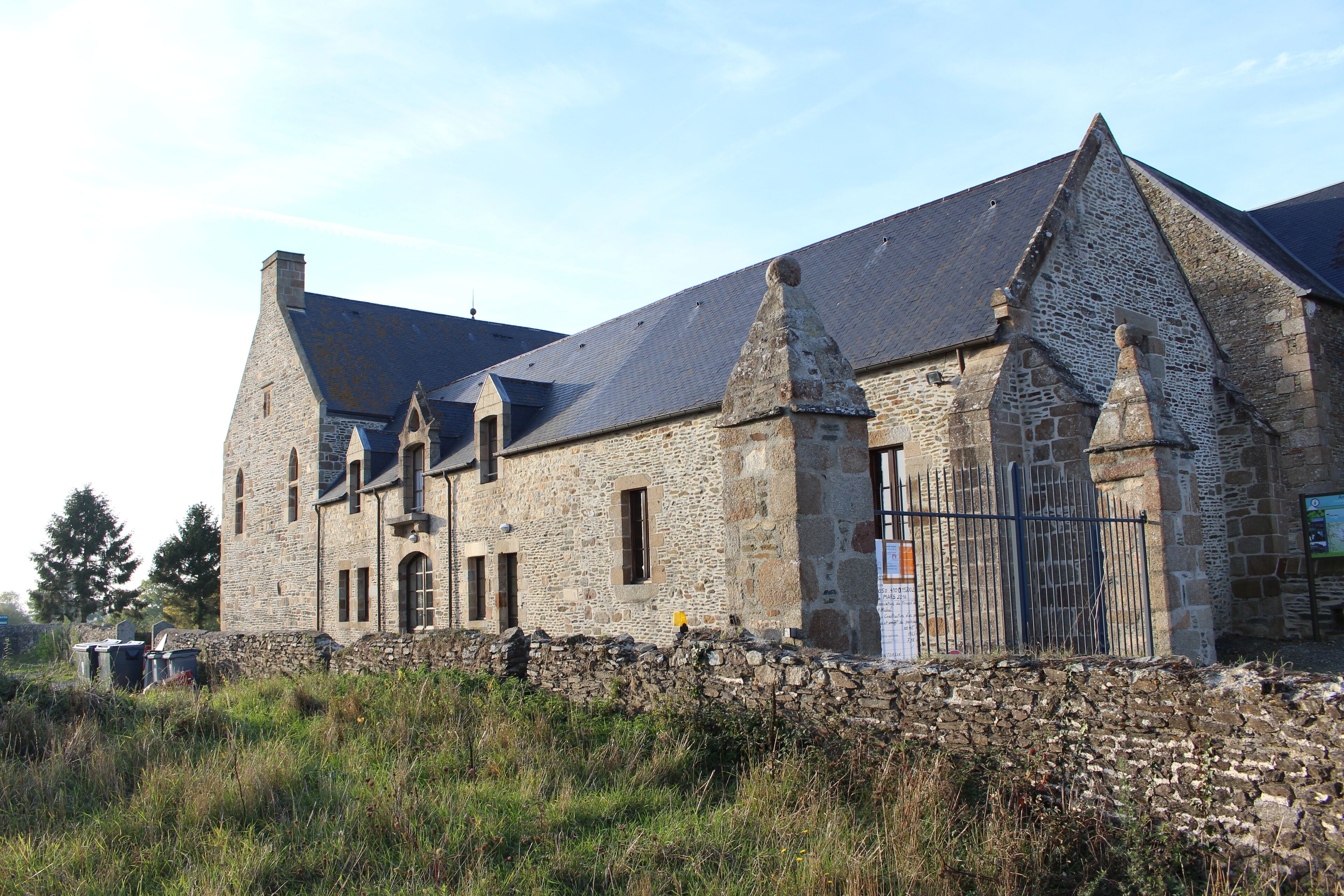 A stone building with a steep roof and a stone wall in front.