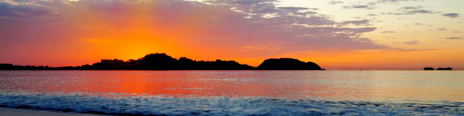 A sunset over a beach with waves, a distant island, and a partly cloudy sky.
