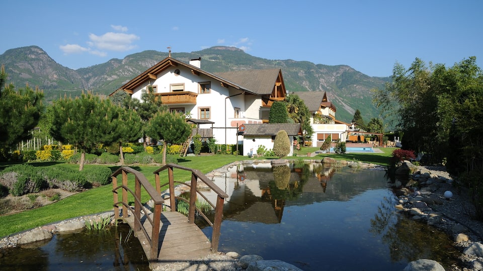 A house with a pond, wooden bridge, and mountains in the background.