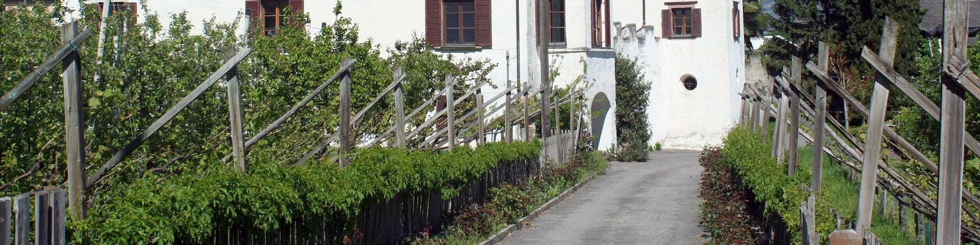 A white building with a red-tiled roof and a bell tower, surrounded by greenery and mountains in the background.