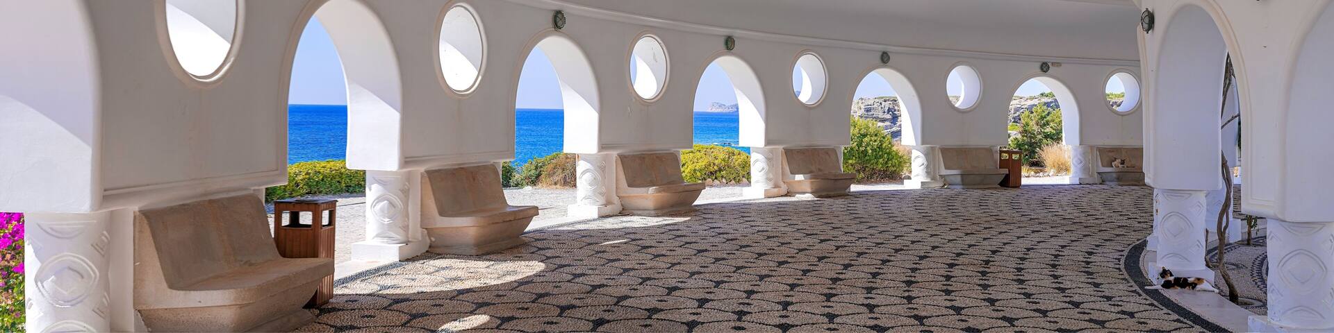A covered outdoor area with white columns and arches, a patterned floor, and a view of the sea.