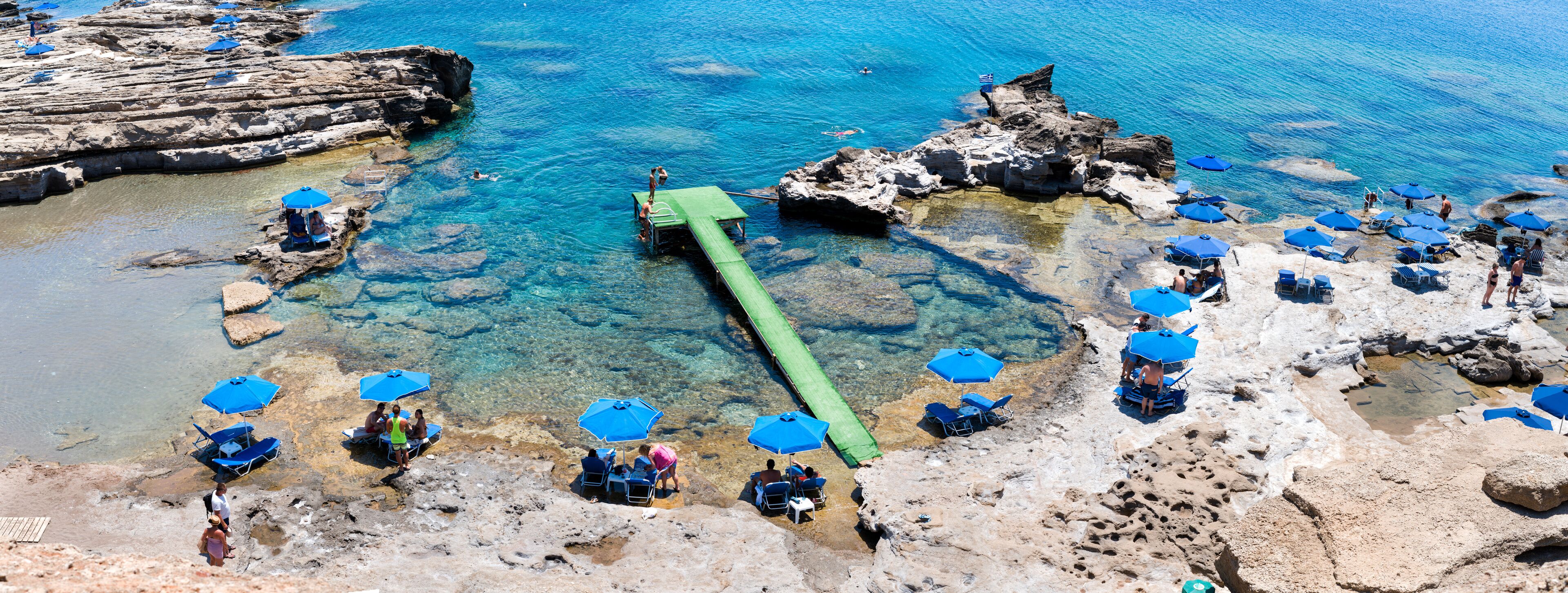 A rocky beach with clear blue water, a green floating walkway, and blue umbrellas.